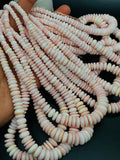 May include: A strand of pink and white shell beads. The beads are round and flat, and they are strung on a thin thread. The beads are arranged in a loose pile, and they are illuminated by a bright light source.